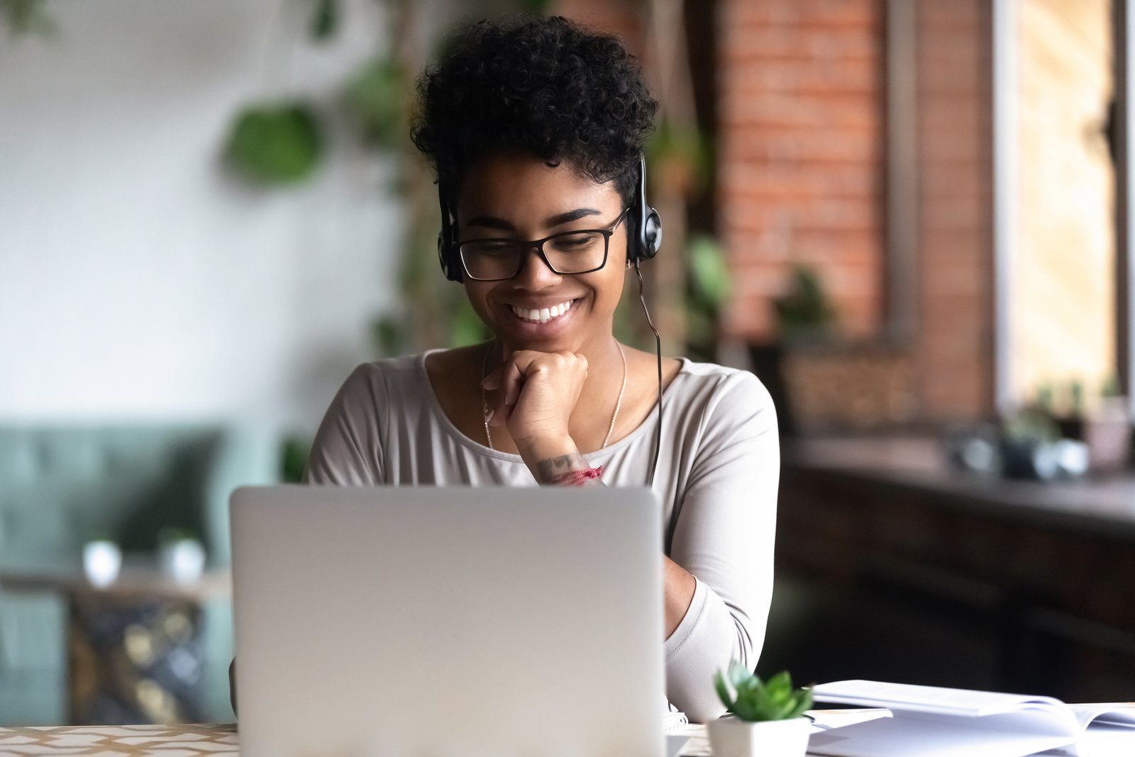 Black woman  sitting at table in university cafe alone wearing glasses looking at computer screen using headphones listening online lecture improve language skills having good mood